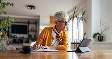 woman at table taking notes while looking at tablet